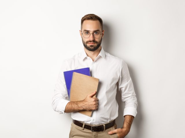 Handsome male employee holding notebooks, looking confident, standing over white background
